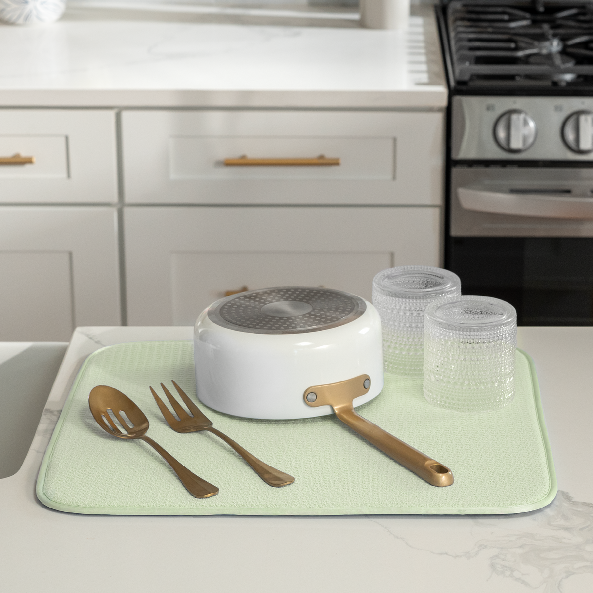A mint green dish drying mat on a white countertop, holding a white pot with a gold handle, two clear textured glasses, and two gold utensils. A white kitchen cabinet and a black gas stove are visible in the background.