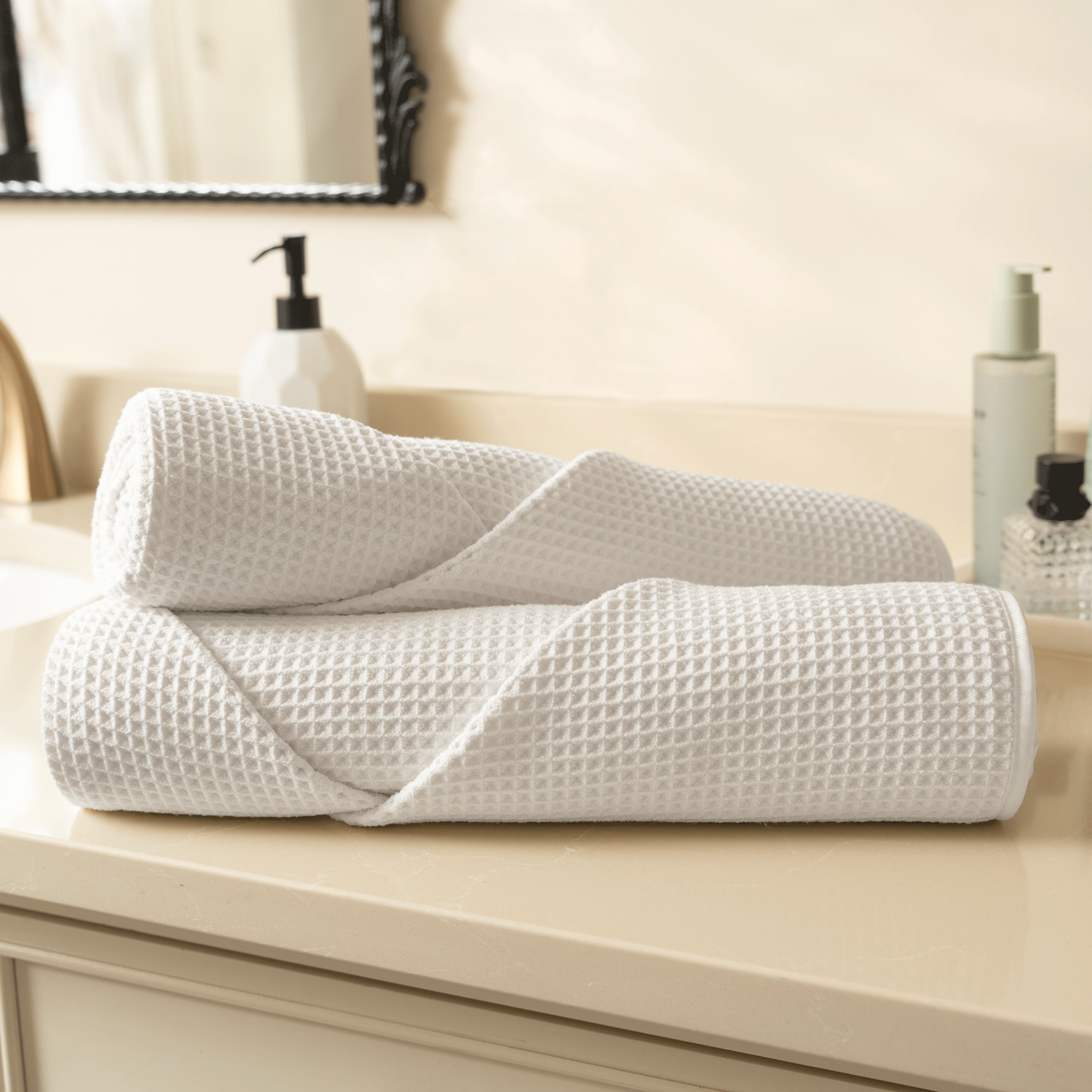 Two rolled white waffle bath towels on a bathroom counter with toiletries in the background.