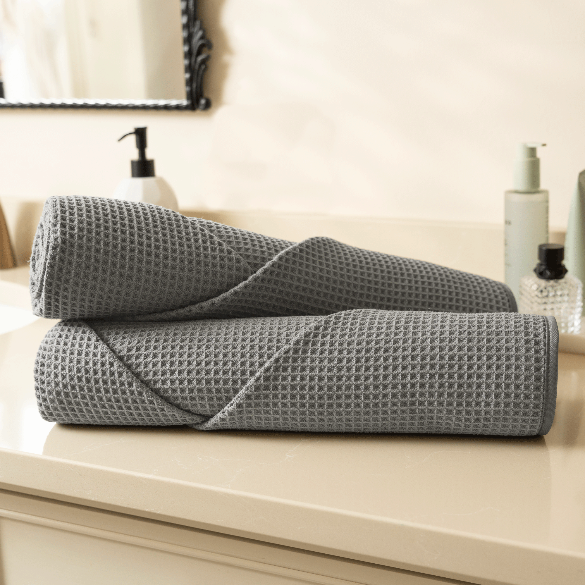 Two rolled grey waffle bath towels on a bathroom counter with toiletries in the background.