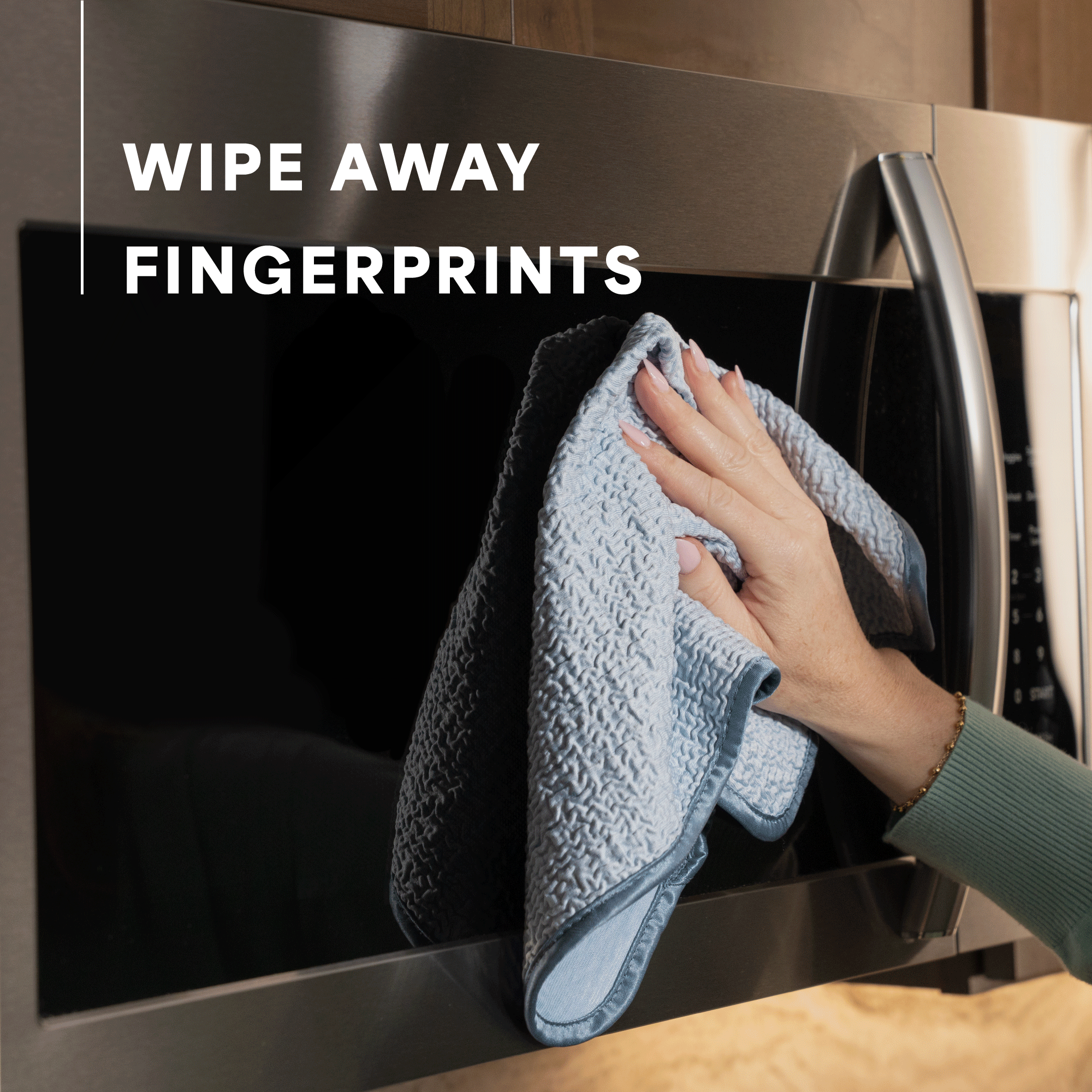A person wipes a stainless steel microwave door with a textured light blue cloth. White text on the left reads 'WIPE AWAY FINGERPRINTS'.