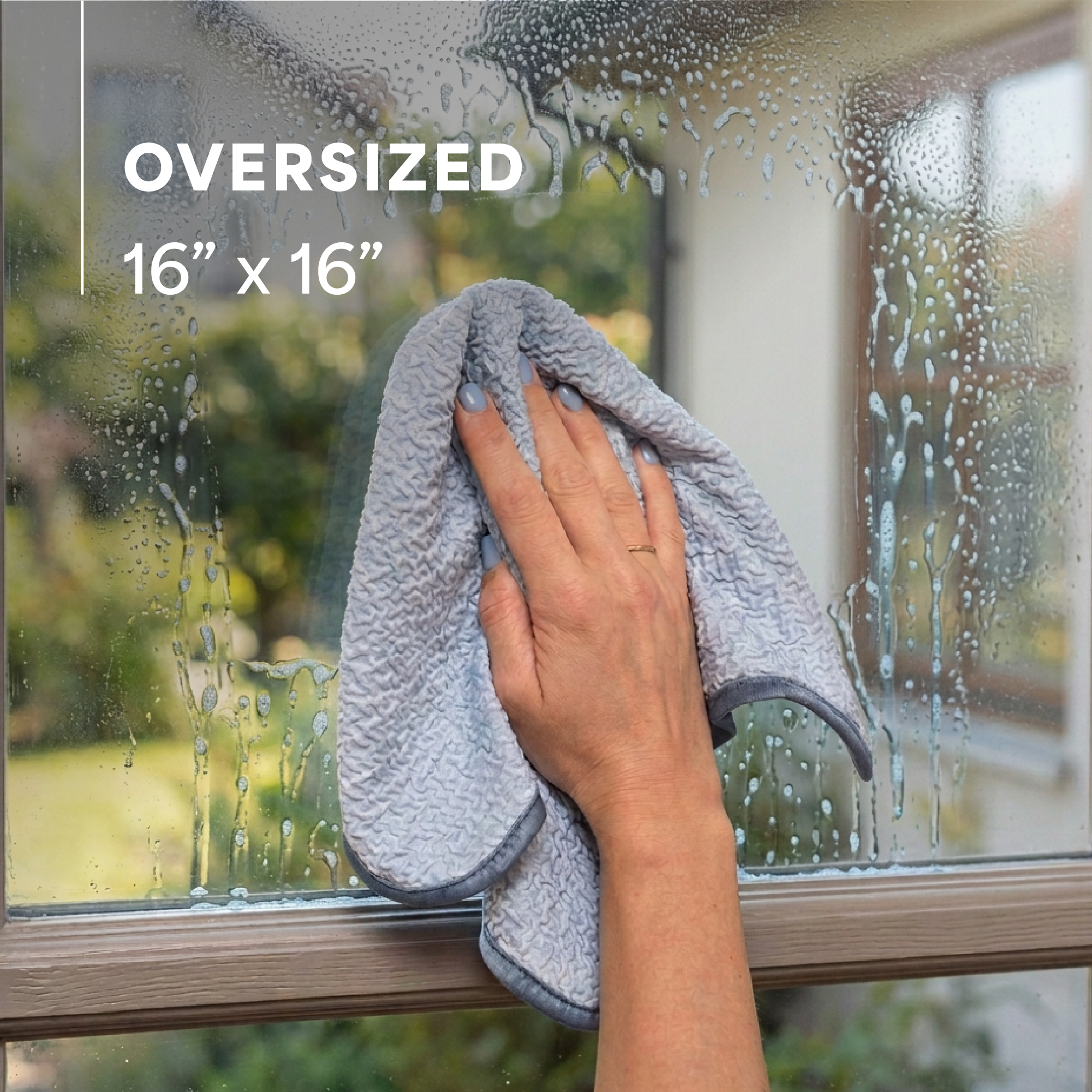 A person wipes a wet, water-droplet-covered window with a light blue microfiber cloth. White text in the upper left reads 'OVERSIZED 16" x 16"'.