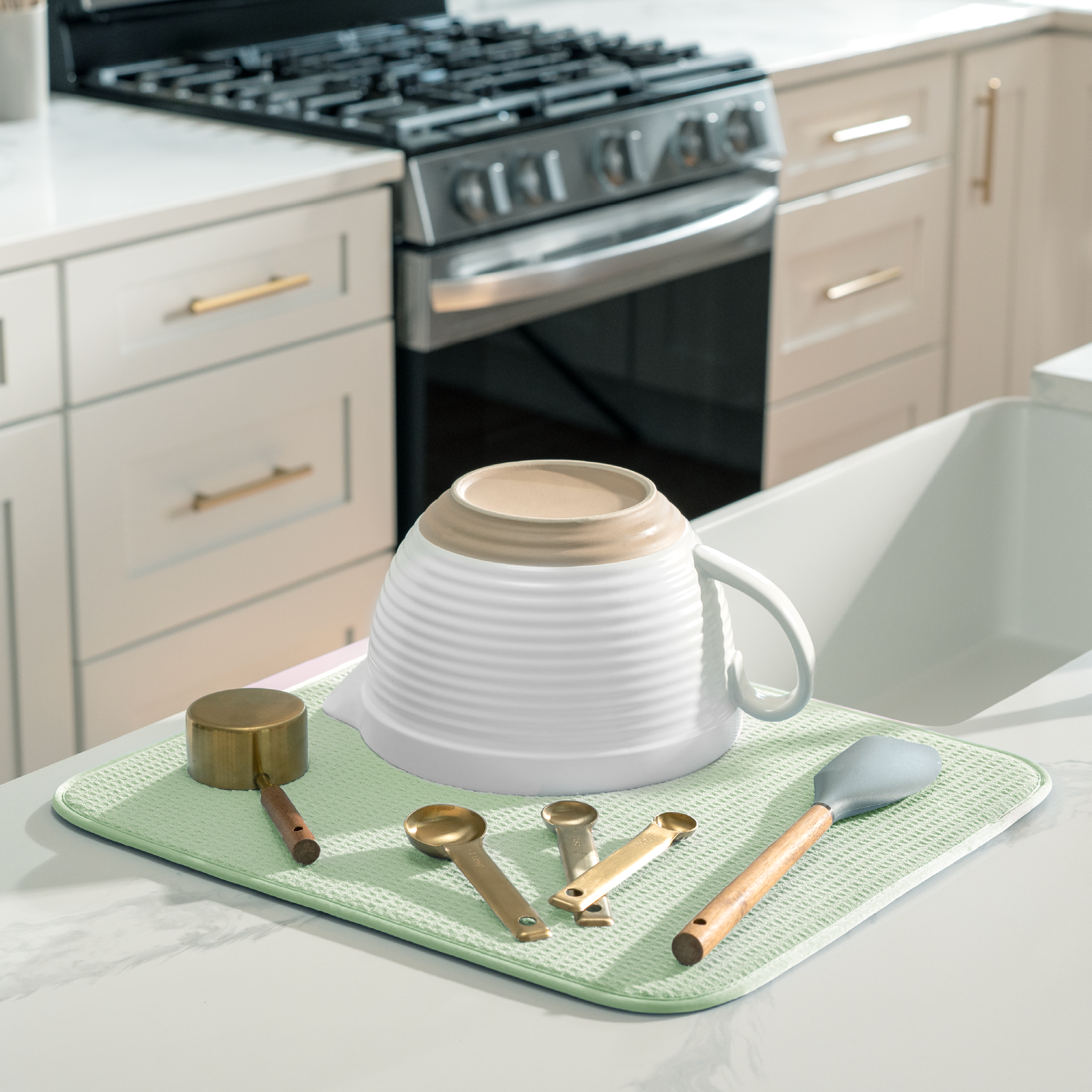 A light green microfiber dish drying mat on a white marble countertop in a modern kitchen setting. The mat is styled with a white ribbed ceramic mug, a gold measuring cup, spoons, and a spatula.