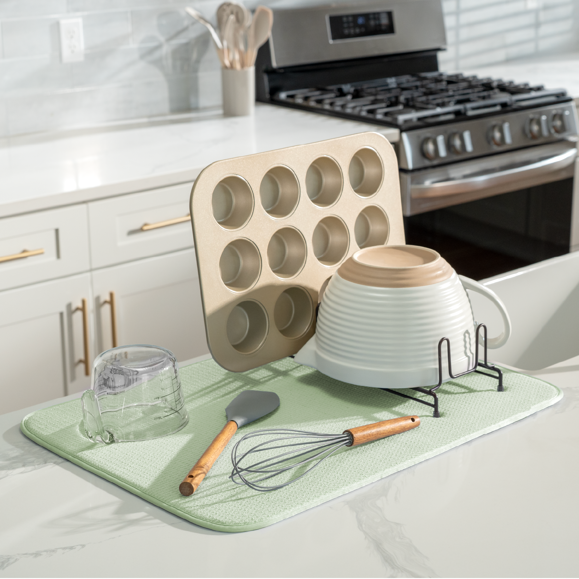 A kitchen scene showing a light green microfiber drying mat on a white countertop, with a  muffin tin, stacked white bowls, a glass, a spatula, and a wire whisk resting on it. A stove and cabinets are visible in the background.